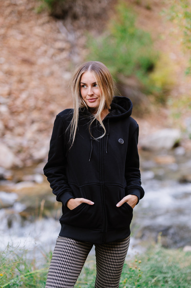Woman wearing a black hoodie and patterned pants standing outdoors near a stream with foliage in the background.