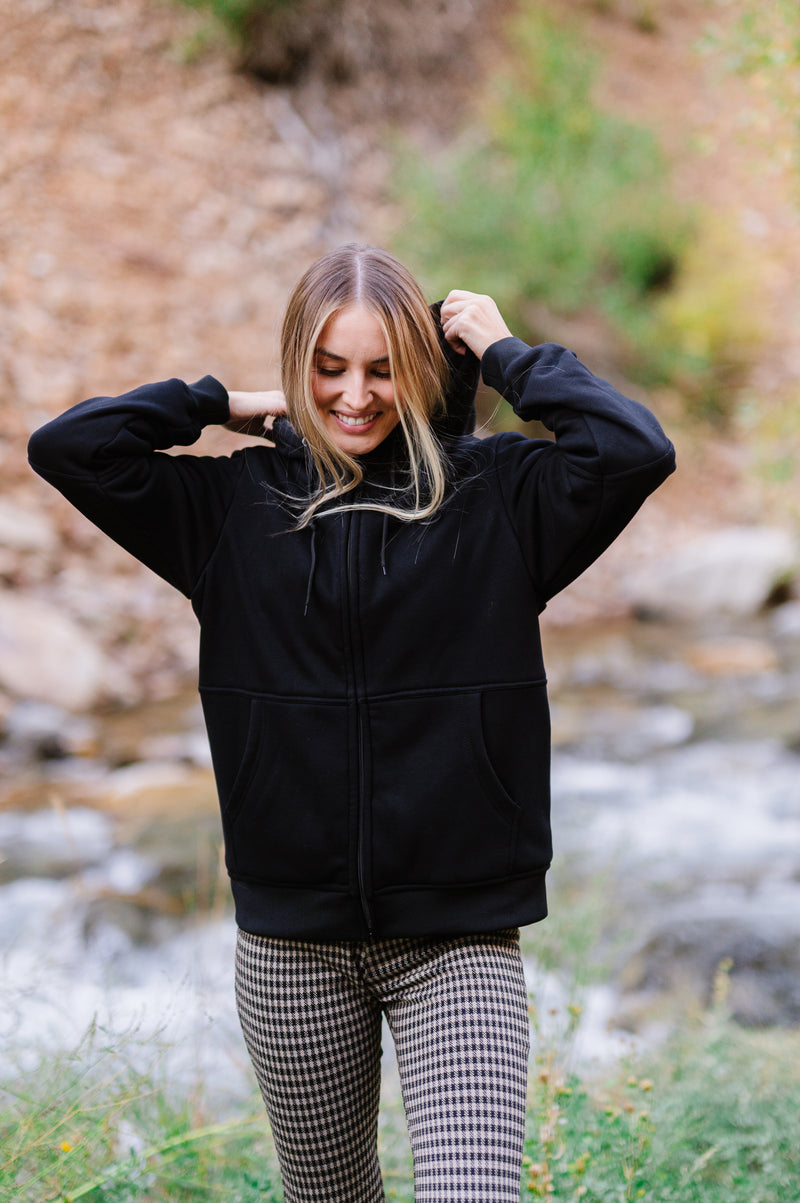 Woman smiling while adjusting hood on black jacket in nature setting with rocky background and flowing stream