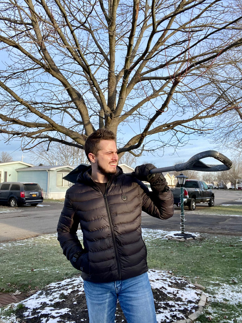 A man in a black winter jacket and blue jeans holding a snow shovel outdoors, standing in a snowy suburban neighborhood with a leafless tree and cars in the background.
