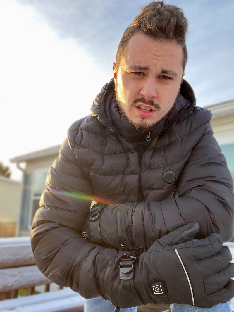 A person wearing a black winter jacket and gloves standing outside on a sunny cold day with a building in the background.