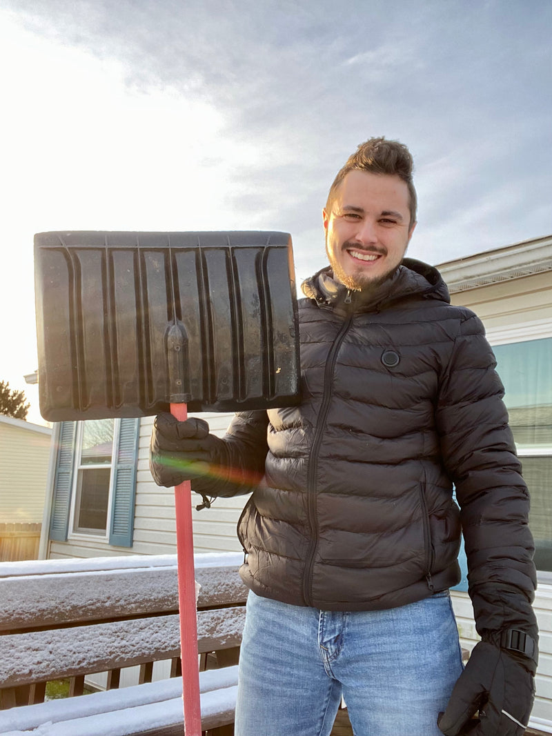 Man smiling while holding a snow shovel on a snowy day, wearing a black winter jacket and gloves.