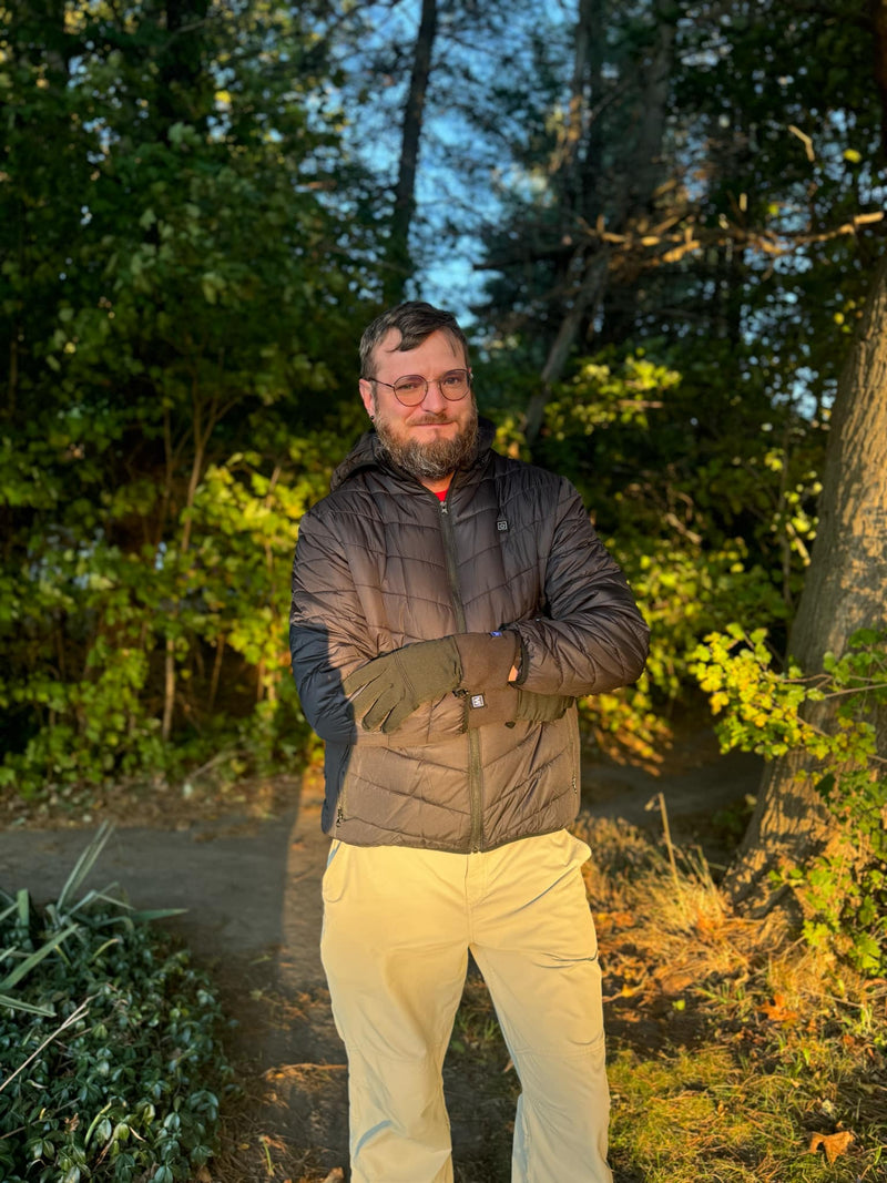 A person wearing glasses and a black jacket standing in a sunlit forest.