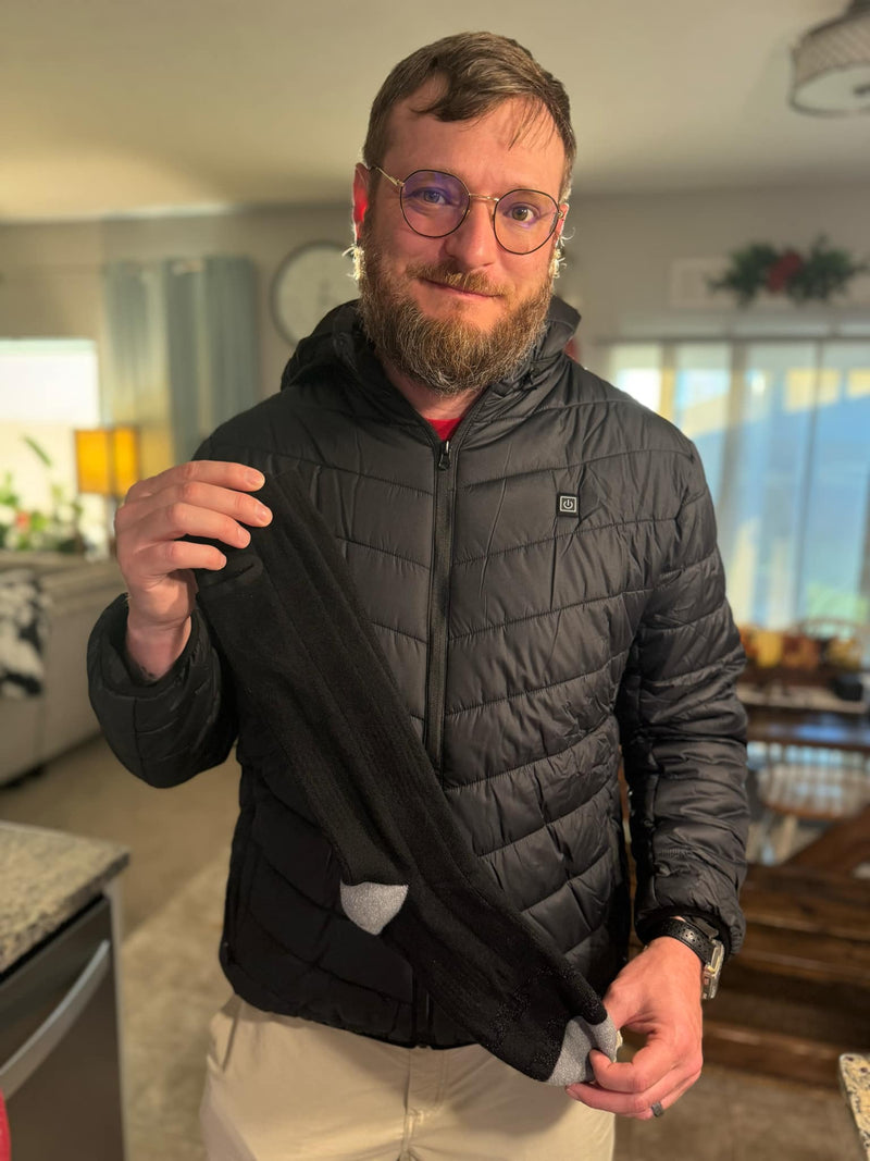 A man with a beard and glasses wearing a black jacket holds a black and gray long sock in a warmly lit living room.