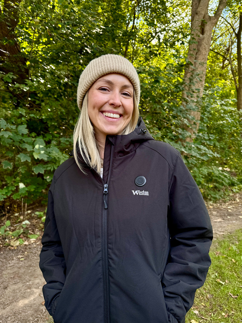 Smiling person wearing a black Weston jacket and beige knit hat with a forest background.