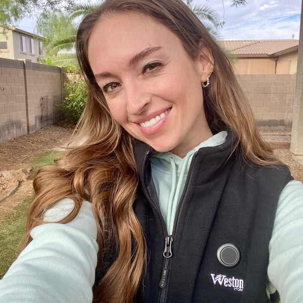 Woman smiling outdoors wearing a black vest with Weston Store logo, with long hair, in a backyard setting with trees in the background.