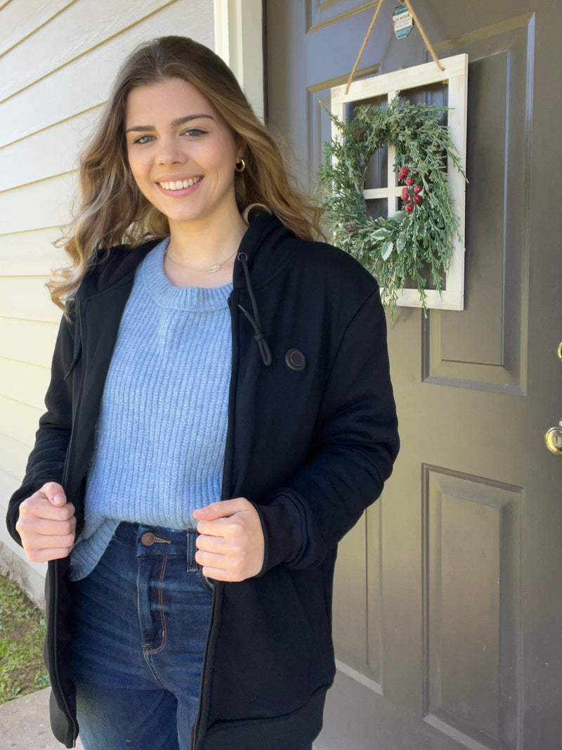 Young woman smiling in front of a door with a decorative wreath, wearing a blue sweater and black jacket.