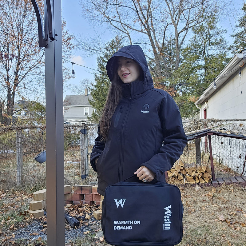 Person wearing a black hooded jacket outside holding a case with "Warmth on Demand" text in a backyard with autumn leaves.