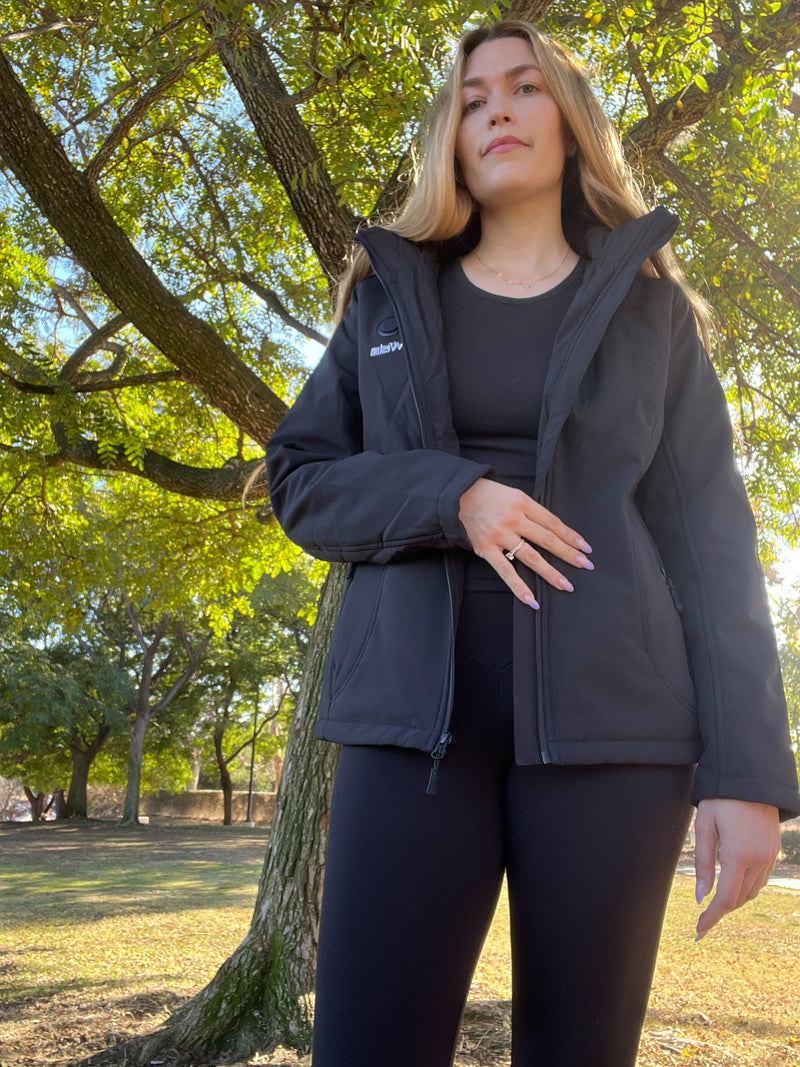 A person wearing a black jacket and leggings stands outdoors near a tree with green leaves, looking confident on a sunny day.