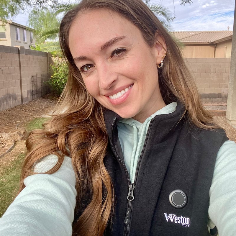 Smiling woman taking a selfie outdoors, wearing a black vest and light green hoodie, with a backyard and brick wall in the background.