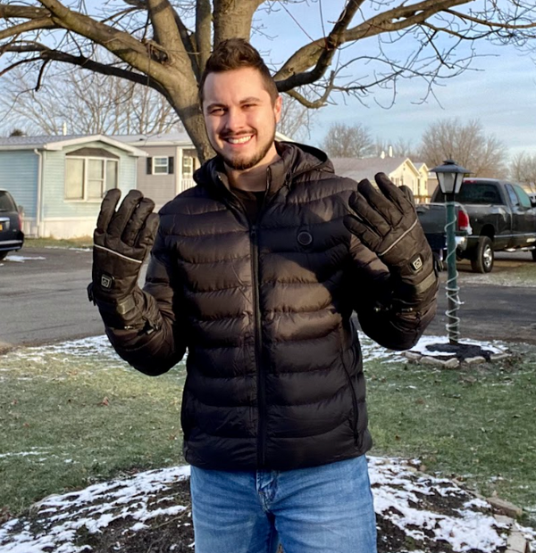 Person smiling and wearing a puffy black jacket with gloves, standing outdoors on a snowy day with trees and houses in the background.