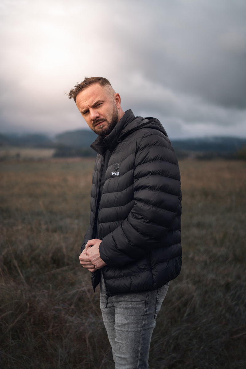 Man wearing a black puffer jacket standing in a grassy field under a cloudy sky.