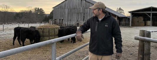 Man in a black Weston heated coat leaning on a frosty cattle gate with a feed bucket during morning chores