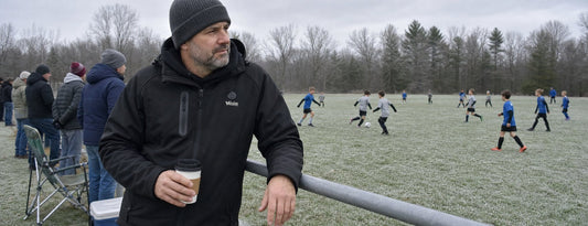 Man in a black Weston heated coat leaning on the sideline rail at a cold youth soccer game