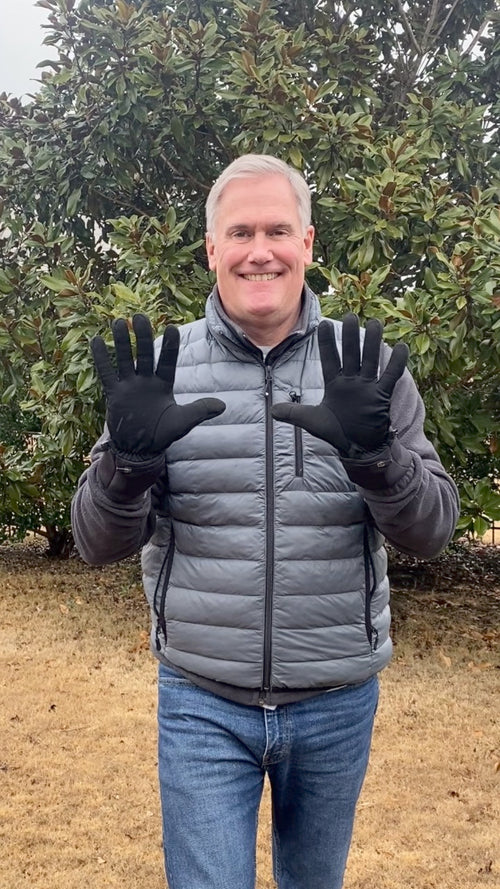 Man wearing a gray puffer jacket and black gloves smiling outdoors in front of leafy green trees.