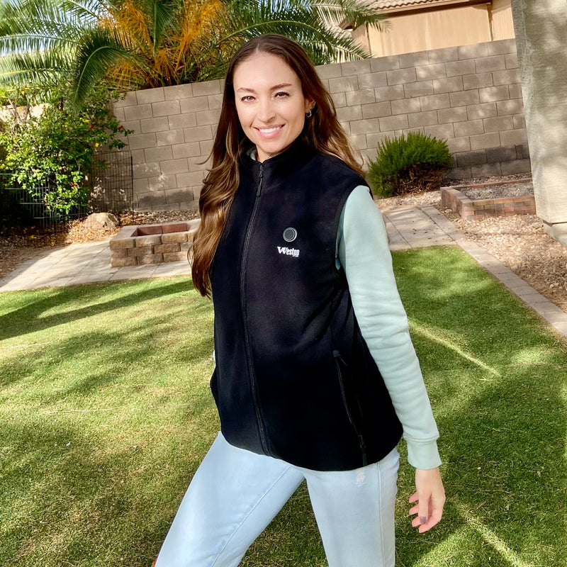 Person in black vest and light blue jeans standing on a green lawn with palm trees and brick wall in the background, smiling.