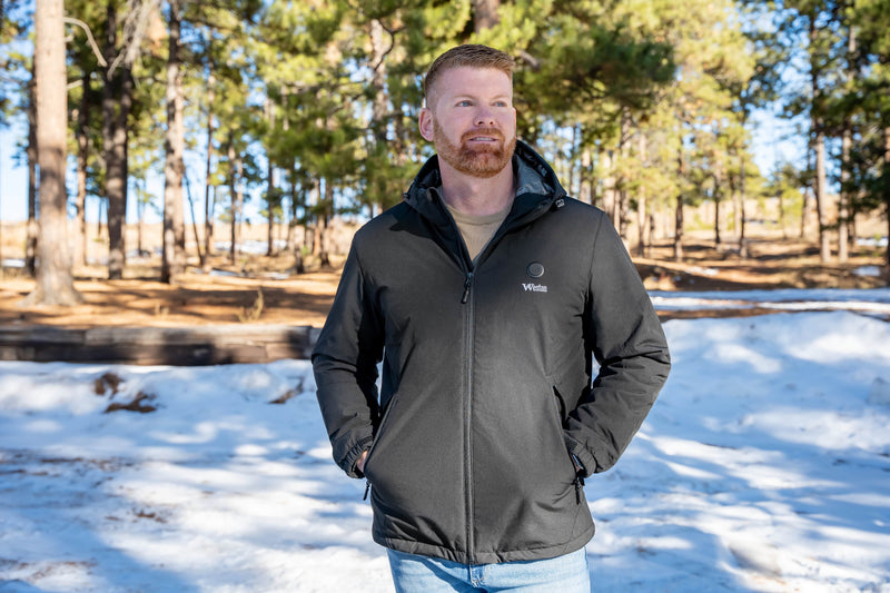 Man wearing a black winter jacket with a beard, standing in a snowy forest setting under sunlight.