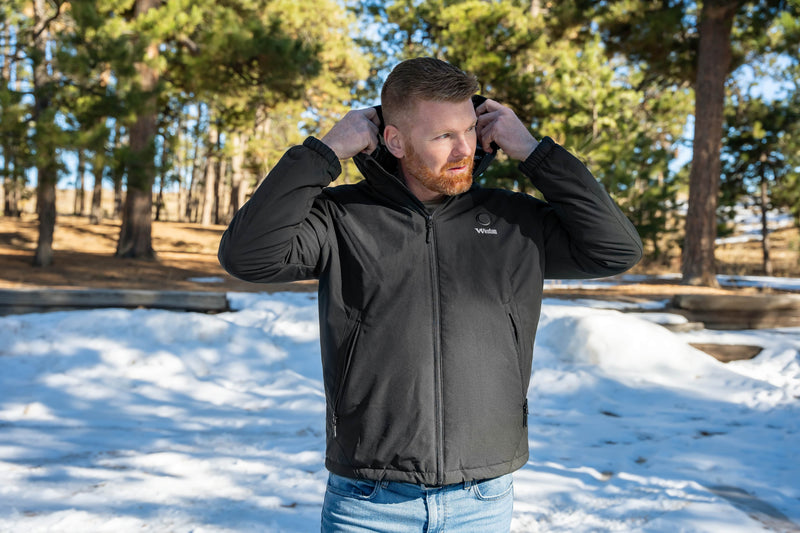 Man wearing a black jacket with hood in snowy forest setting.