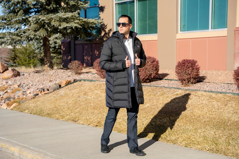 Man wearing a black puffer coat and sunglasses standing on a sidewalk in front of a building with shrubs.