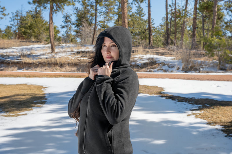 Woman in black hoodie standing outdoors in a snowy landscape with evergreen trees in the background.