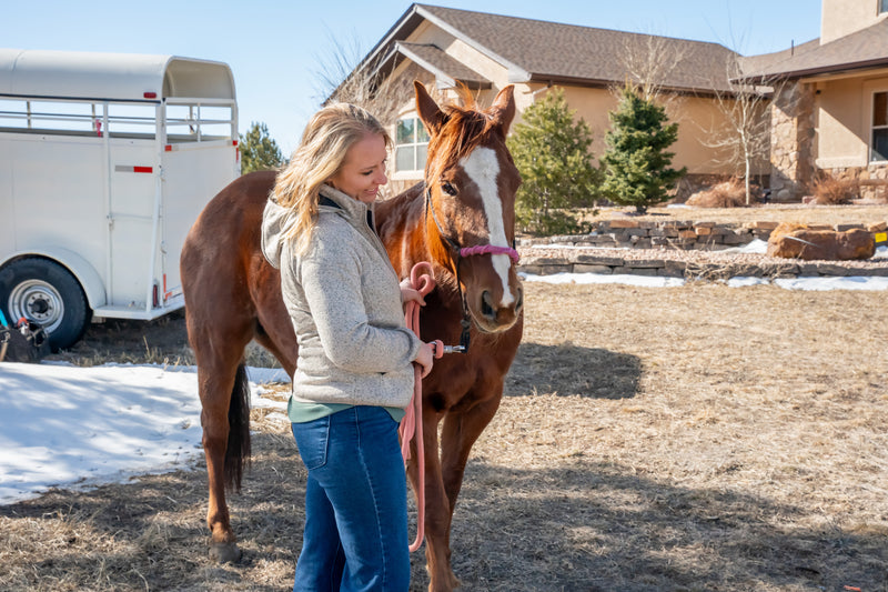 Woman standing outdoors with a brown horse, holding a pink lead rope next to a horse trailer and house.