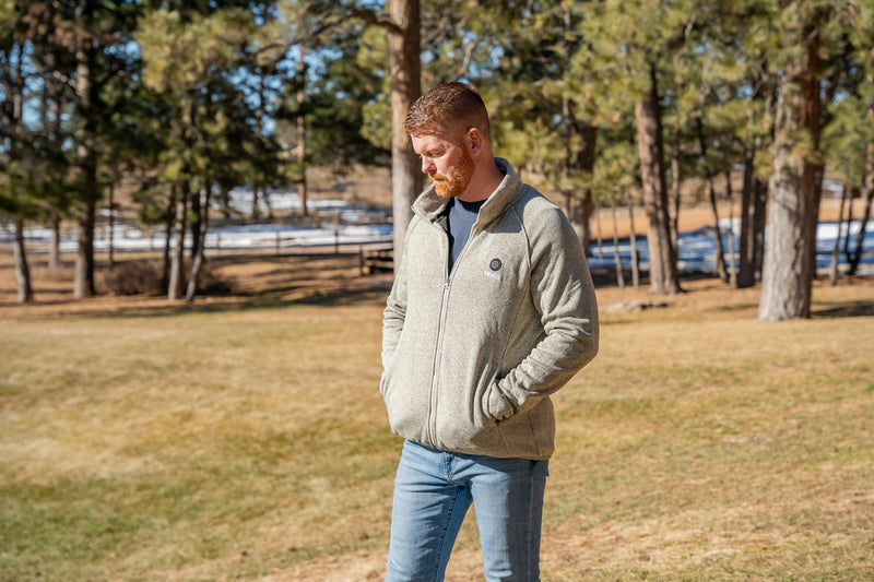 Man wearing a gray fleece jacket and blue jeans standing outdoors in a park with trees in the background.