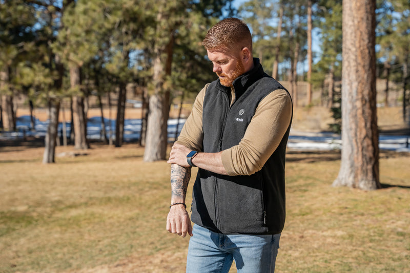 Man adjusting his sleeve in a forest setting wearing a black vest and beige shirt, with tattoos on his arm and a smartwatch on his wrist.