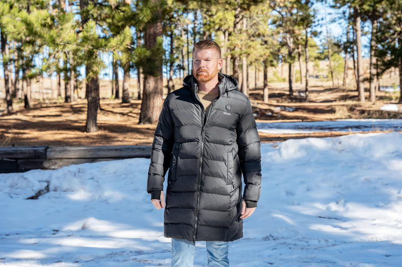 Man wearing a black winter coat standing in a snowy forest setting.