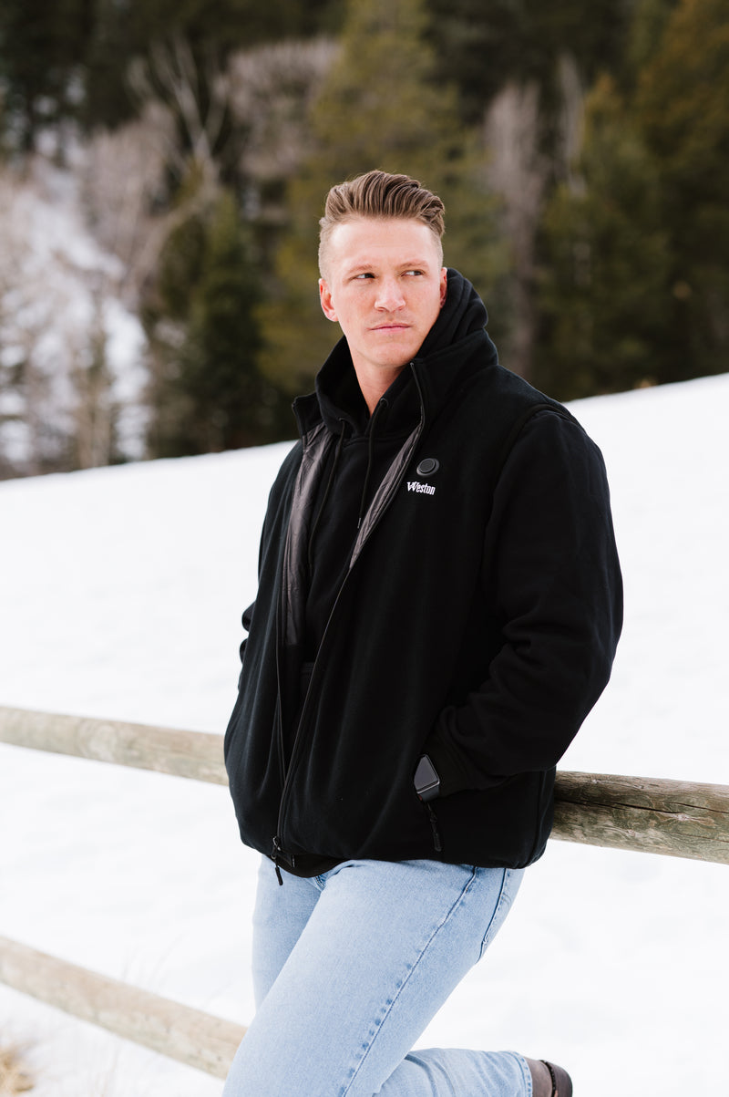 A person in a black jacket and light blue jeans leaning against a wooden fence with a snowy forest background.