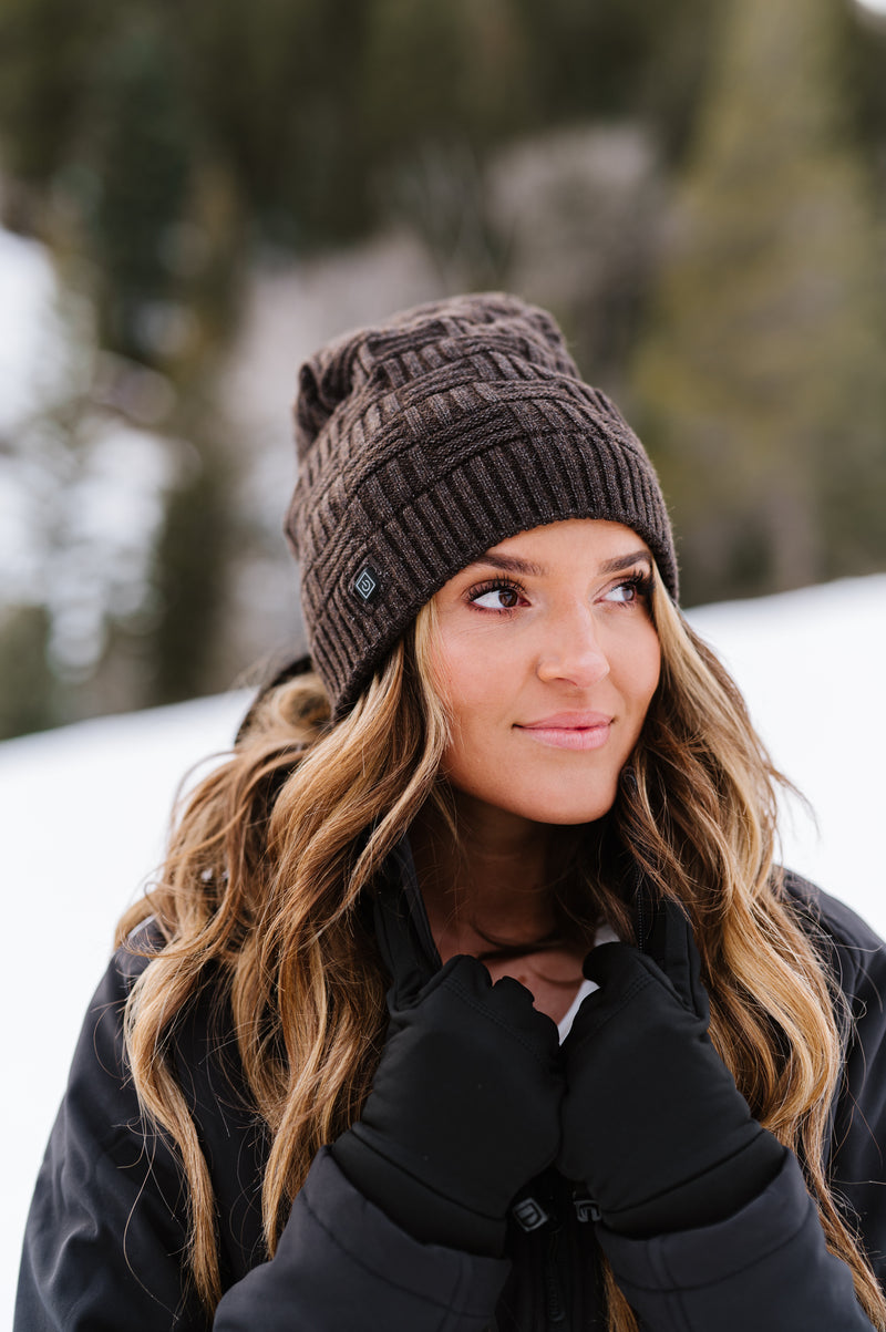 Woman wearing a brown knit beanie and black jacket, standing in a snowy forest with a thoughtful expression.