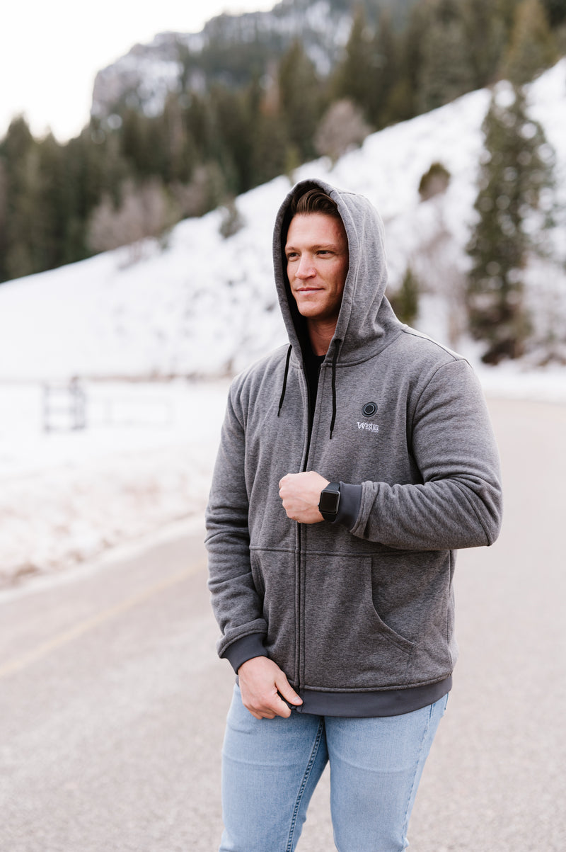 Man wearing a gray hoodie and smartwatch standing on a snowy mountain road with evergreen trees in the background.