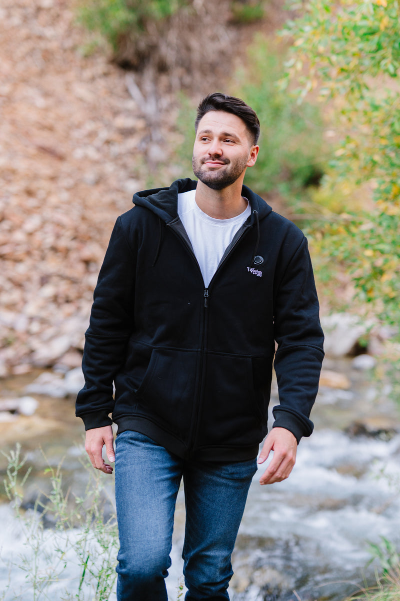 Man in black hoodie and jeans walking near a stream with a blurred natural background