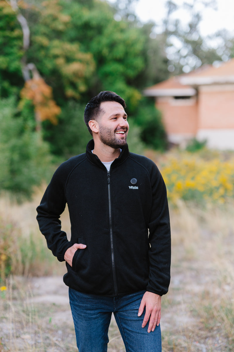 Man in a black jacket standing outdoors in a park with green trees and yellow flowers in the background.
