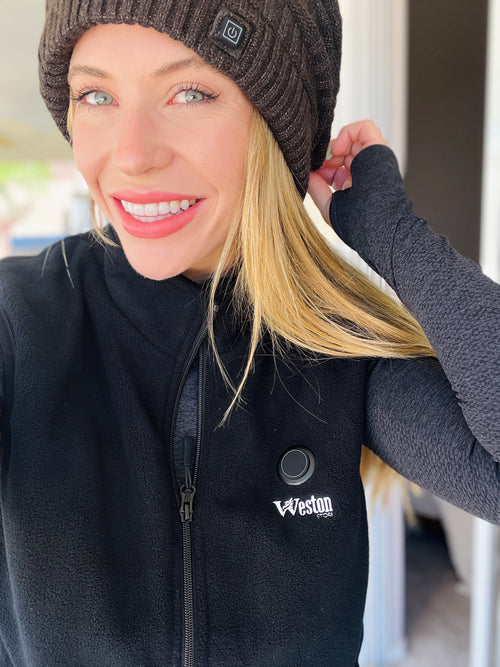 Woman smiling wearing a black fleece jacket and a knitted beanie indoors.