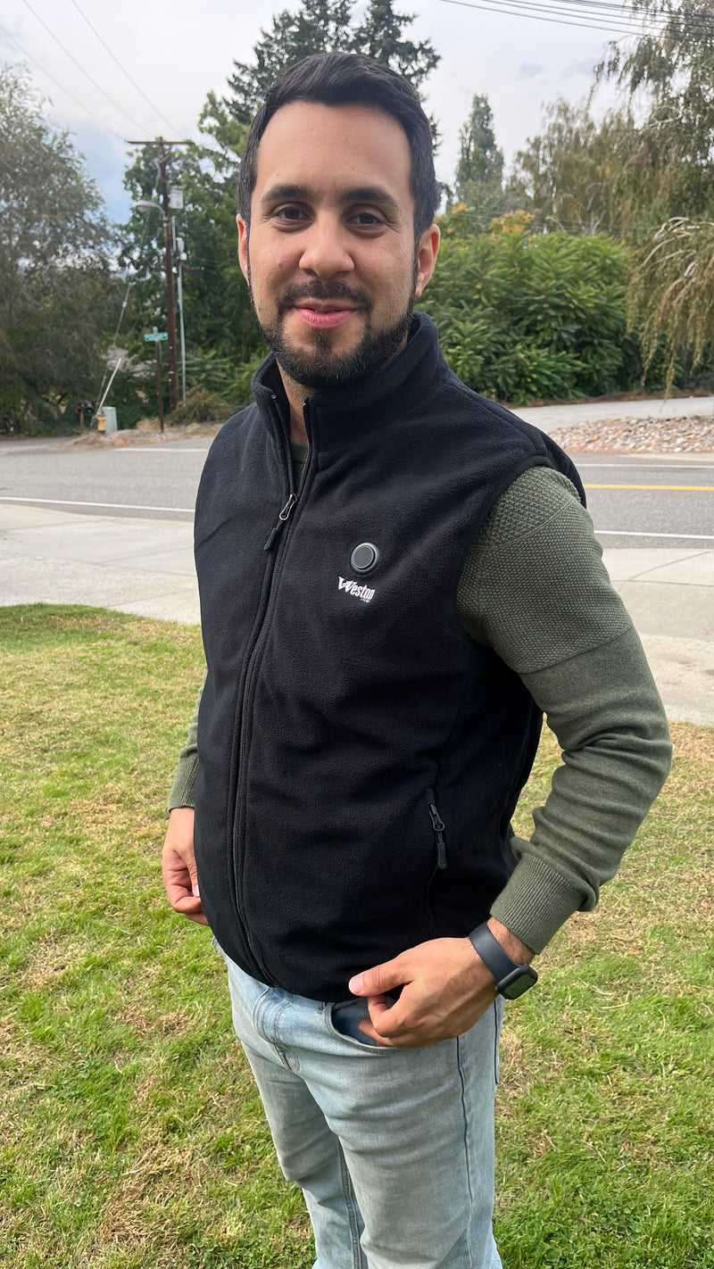 Man wearing a black vest and green sweater standing outdoors on a grassy area near a road.