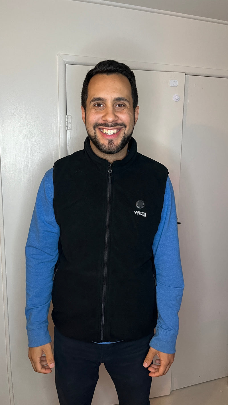 Smiling man in a black vest and blue long sleeve shirt standing indoors.