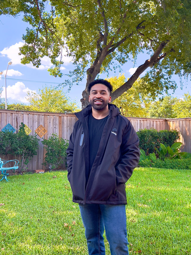 Man smiling in a backyard with green lawn, wearing a black jacket and blue jeans, standing in front of a wooden fence with greenery and a large tree overhead under a blue sky.