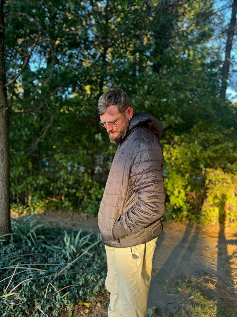 Man in a brown jacket and beige pants standing outdoors surrounded by green trees in sunlight.