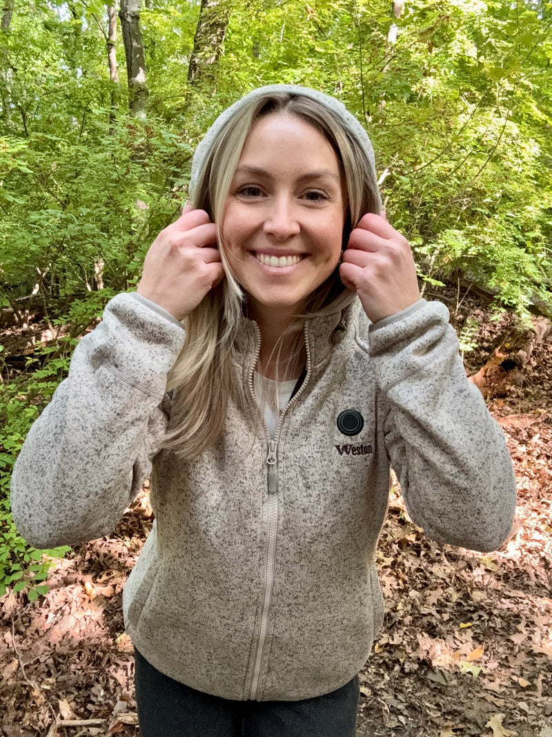 Woman smiling and pulling hoodie strings in a forest setting, wearing a light gray jacket with "Weston" logo.