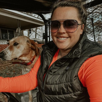Woman wearing sunglasses and black vest smiling with dog beside her outdoors.