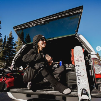 Person sitting in the back of an open SUV surrounded by snowboarding gear with trees and clear blue sky in the background.