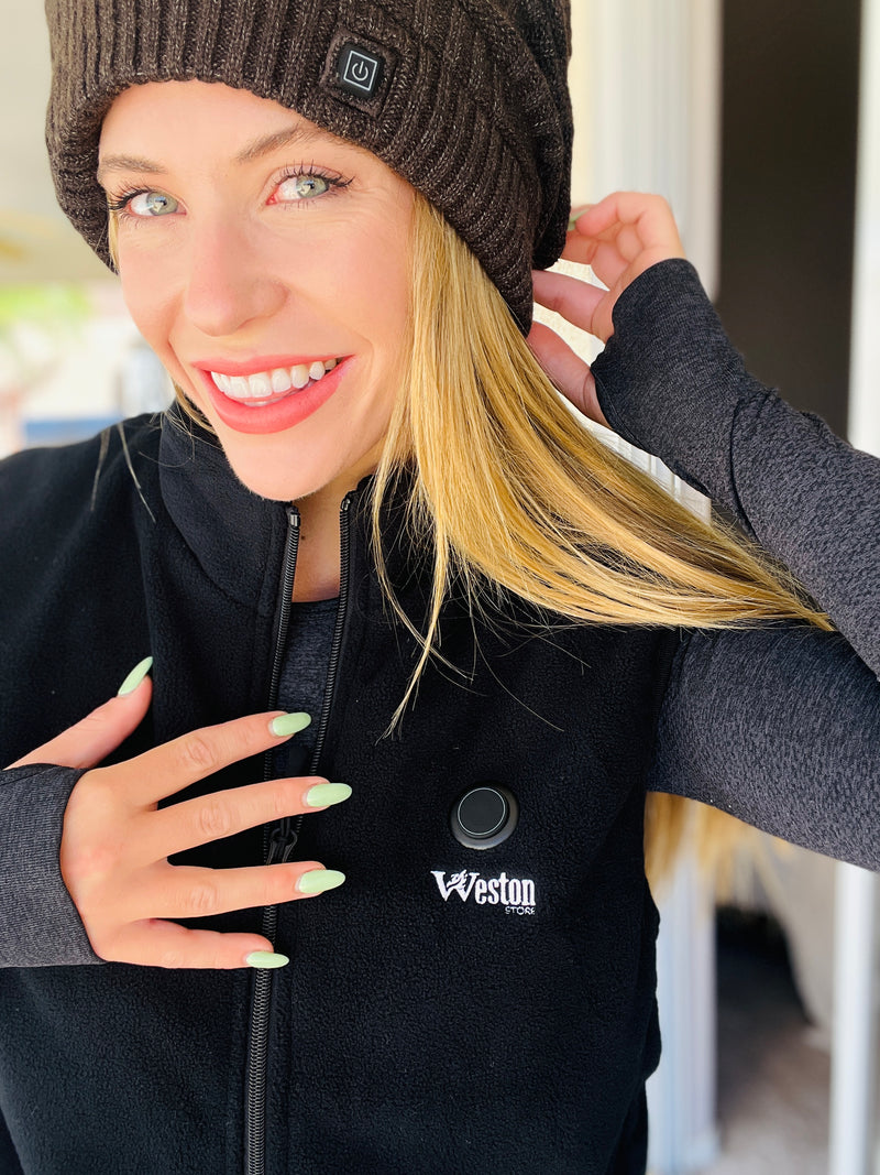 Woman wearing a black fleece vest and a brown knit hat, smiling and holding the hat brim indoors.