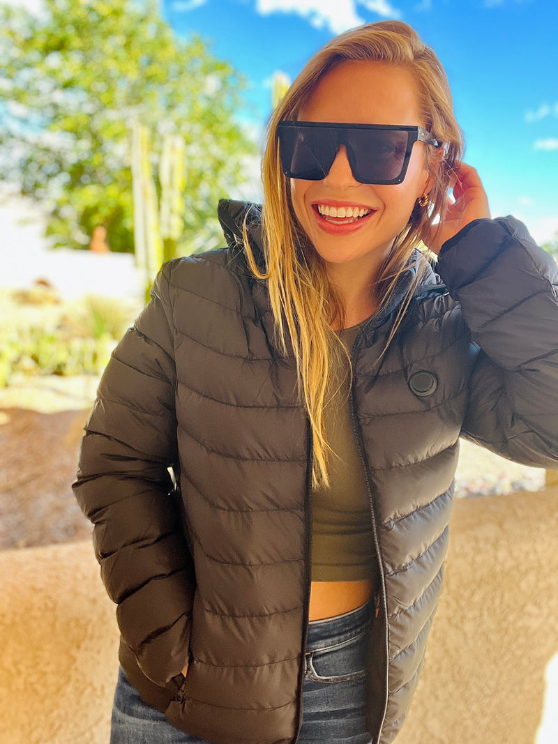 Woman in sunglasses and a black puffer jacket smiles outdoors on a sunny day.
