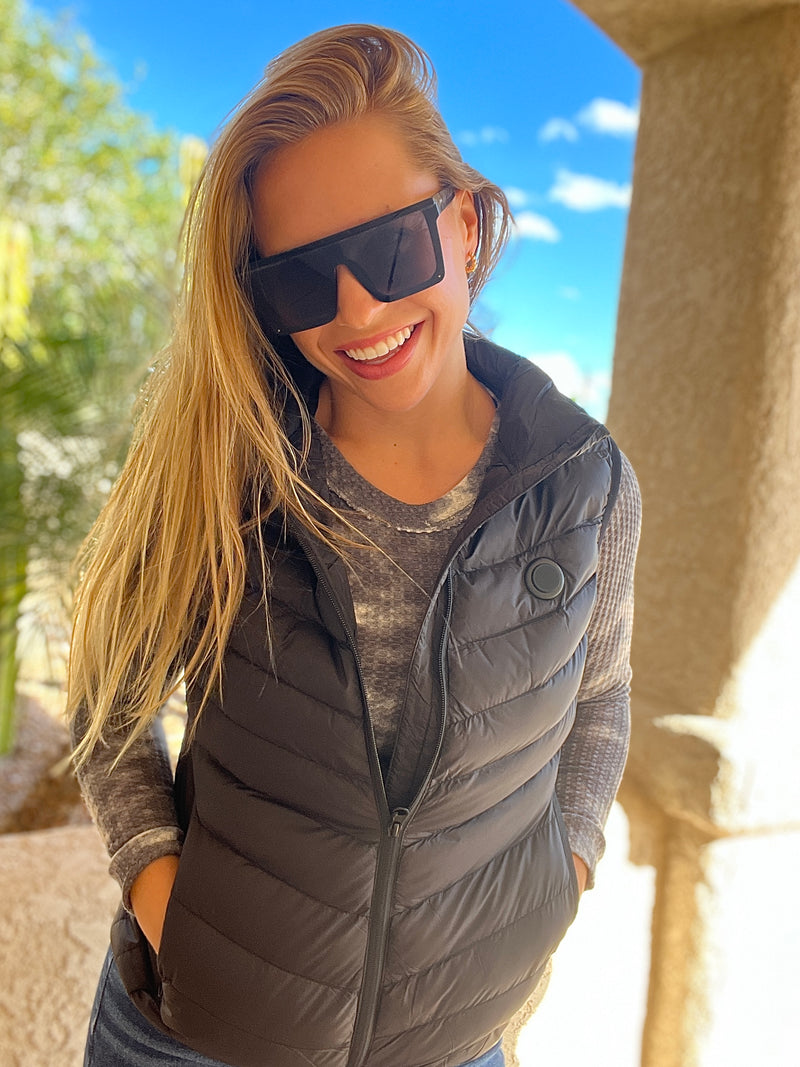 Smiling woman wearing large sunglasses and a black puffer vest, outdoors on a sunny day.