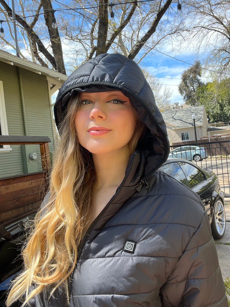 Young woman wearing a black hooded jacket stands outdoors, posing confidently with long wavy hair under a sunny blue sky near a house and parked car.