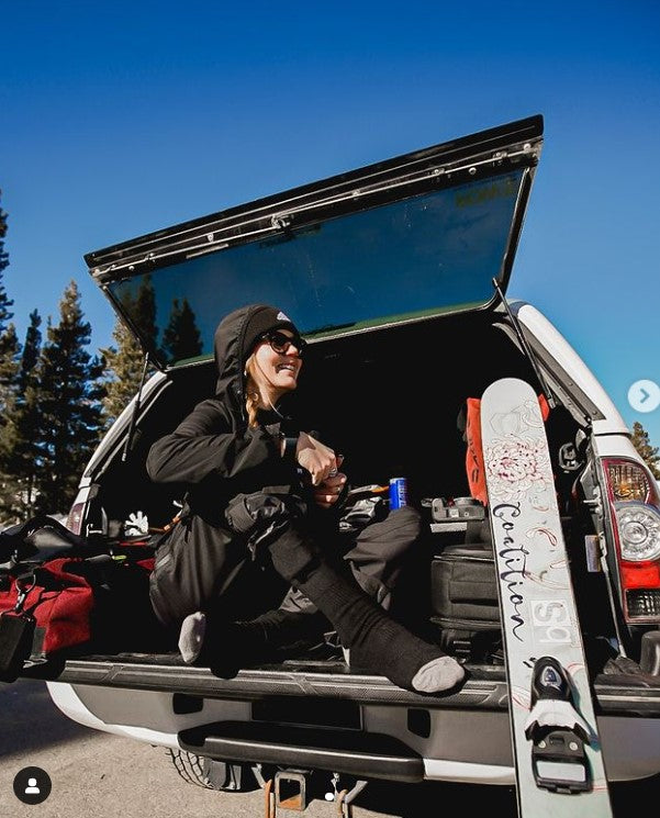 Person sitting in the back of a pickup truck wearing ski gear, with a snowboard and gear visible, enjoying a sunny day with trees and a clear blue sky in the background.