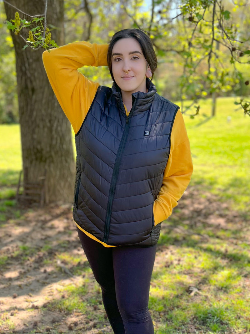 Person in a yellow sweatshirt and black vest posing outdoors in a sunny park with green grass and trees.