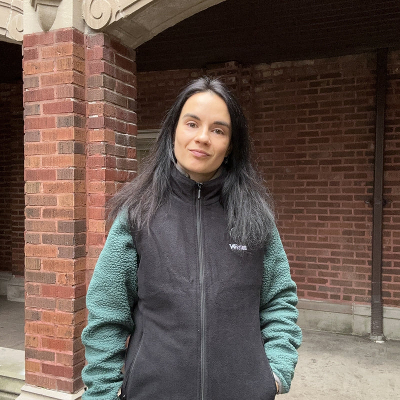 A woman with long dark hair wearing a black and green jacket standing in front of a brick building.