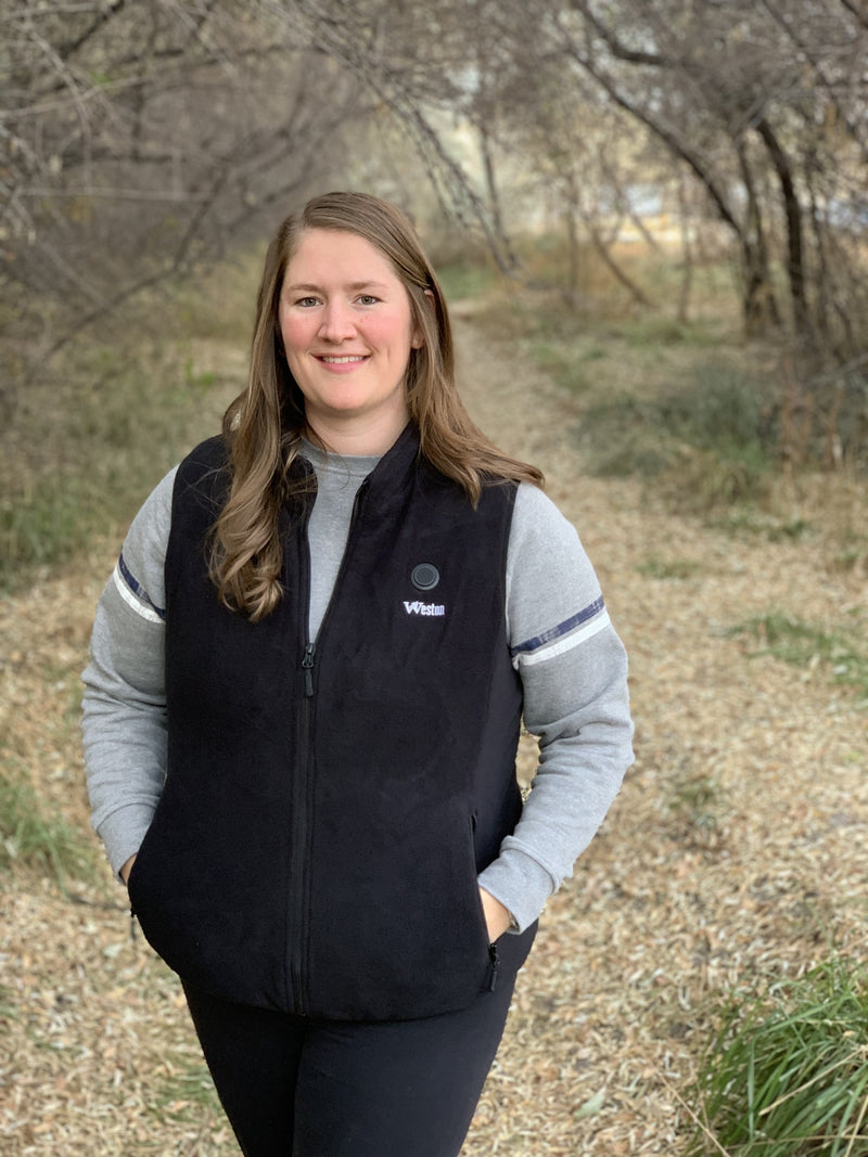 A person wearing a black vest and gray sweatshirt stands on a rustic, wooded path lined with trees and dry grass.