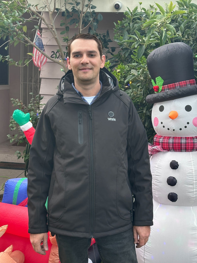 A person wearing a black jacket stands in front of a festive holiday display featuring a large snowman inflatable decoration, with greenery and an American flag in the background.