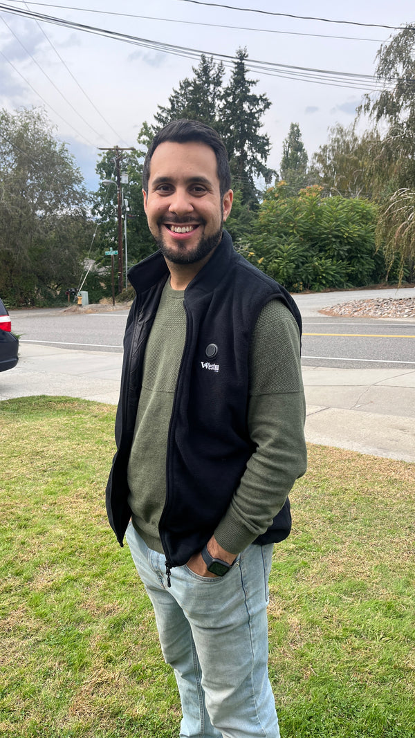 A smiling man with a beard wearing a black vest and green sweater stands outdoors on a grassy area near a street lined with trees and bushes.